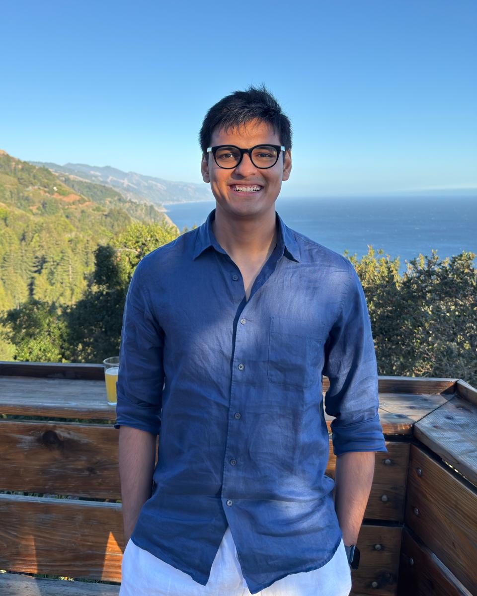 Shivam Raval, founder of Thrivepine, smiling on a wooden deck with a forested coastline behind him